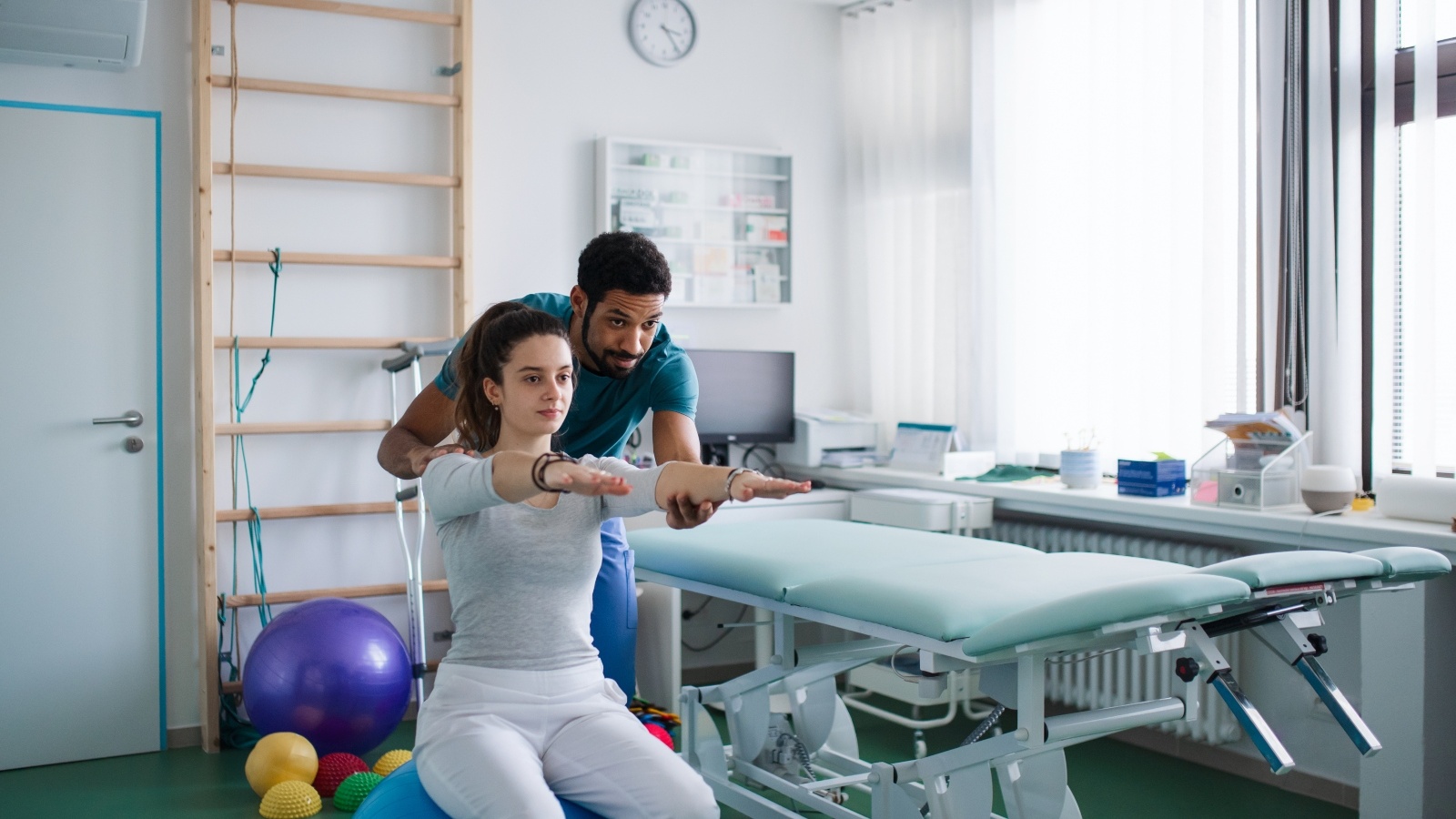 A young male physiotherapist exercising with young woman patient on ball in a physic room.