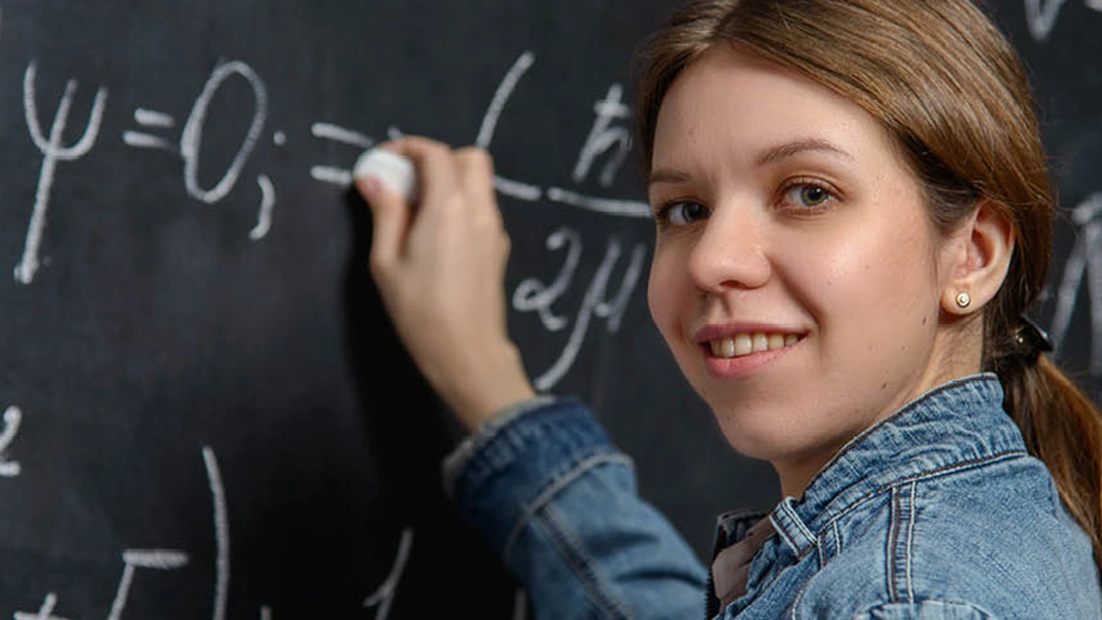 Student solving a physics equation on a black board