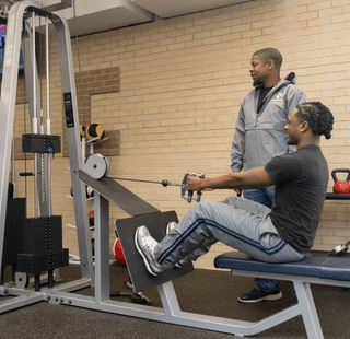 A student lifts weights while another student looks on.