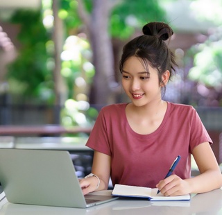 A young woman is happily studying online using her laptop and notebook. She is taking notes and seems to be enjoying her education course in a bright and airy space.