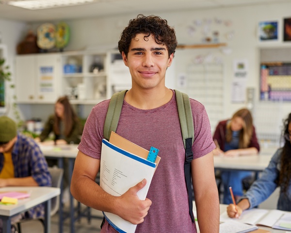 A male high school student holding his books and backpack in a classroom